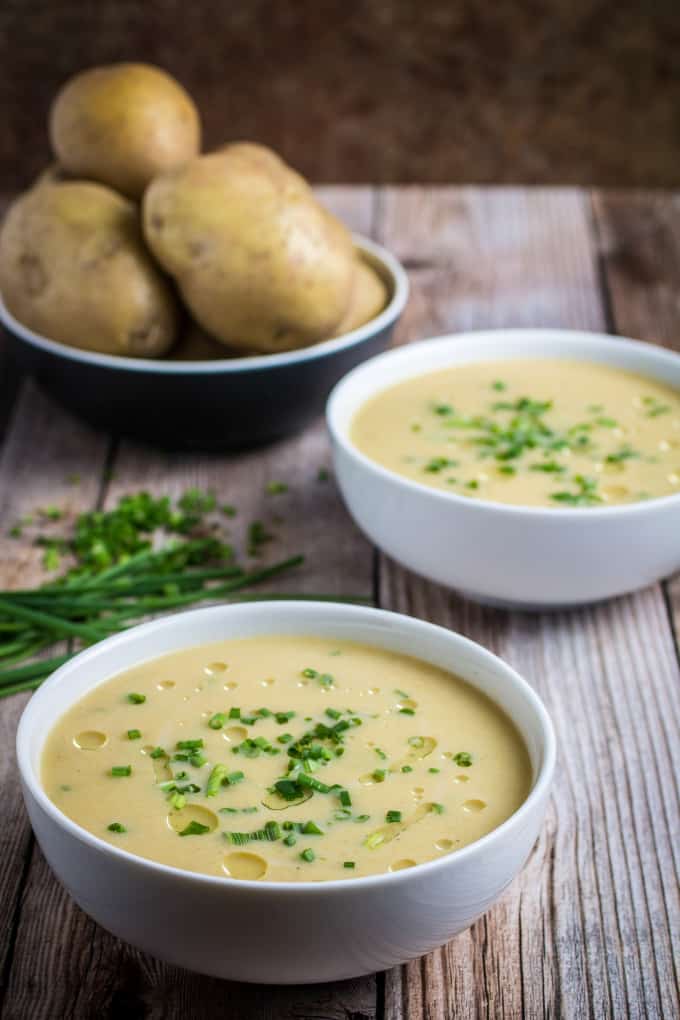 Two bowls of potato leek soup topped with chopped chives on a wooden table, with a bowl of potatoes in the background.