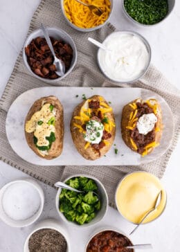 3 baked potatoes on a cutting board with cheese, bacon, and other toppings