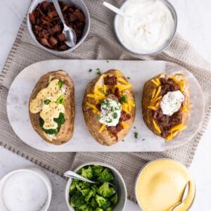 3 baked potatoes on a cutting board with cheese, bacon, and other toppings