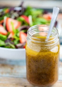 Side view of mason jar full of poppy seed dressing with spoon and strawberry salad in the background