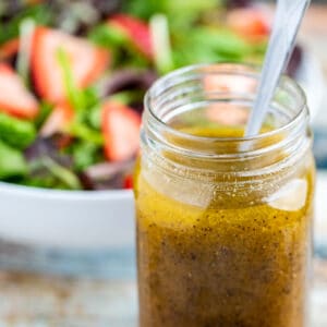 Side view of mason jar full of poppy seed dressing with spoon and strawberry salad in the background