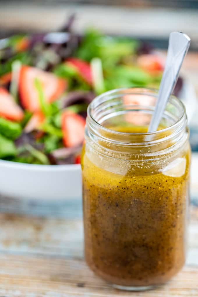 Side view of mason jar full of poppy seed dressing with spoon and strawberry salad in the background