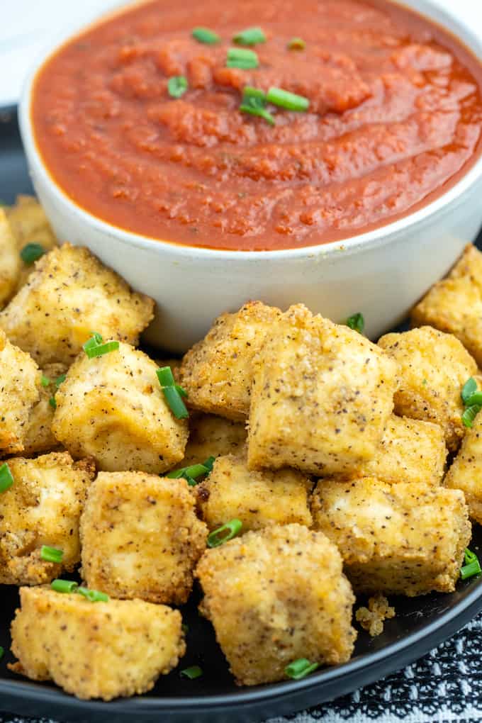 Side photo of air fried golden brown tofu cubes on a plate next to a bowl of marinara dipping sauce