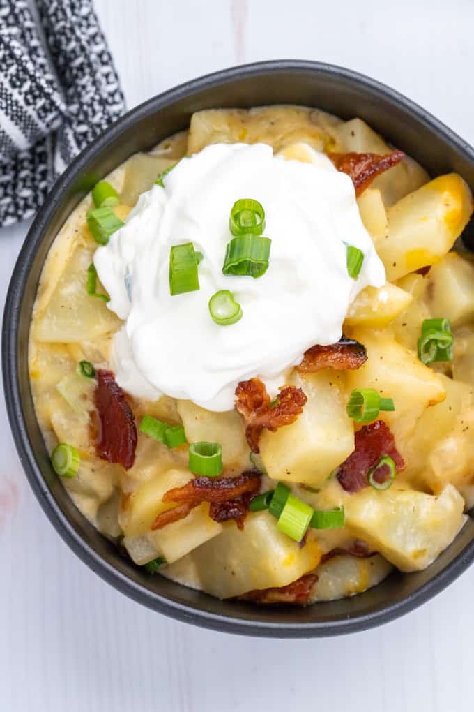 Top down view of a black bowl full of baked potato casserole topped with bacon, green onions, and sour cream.