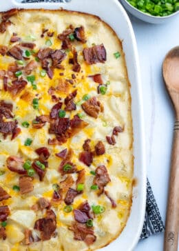 Top down view of casserole dish with loaded baked potato casserole topped with bacon and green onions. Wooden spoon on the table to the right.