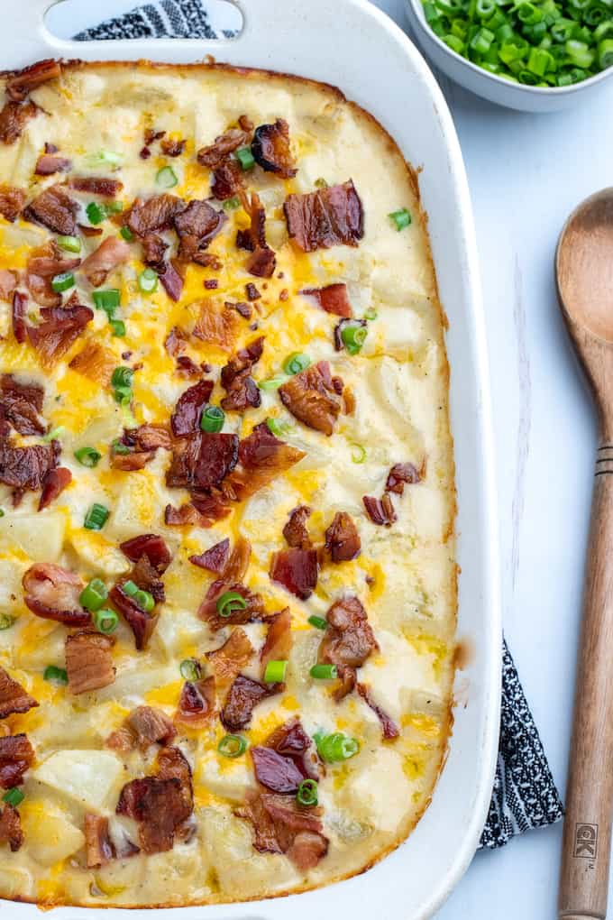Top down view of casserole dish with loaded baked potato casserole topped with bacon and green onions. Wooden spoon on the table to the right. 