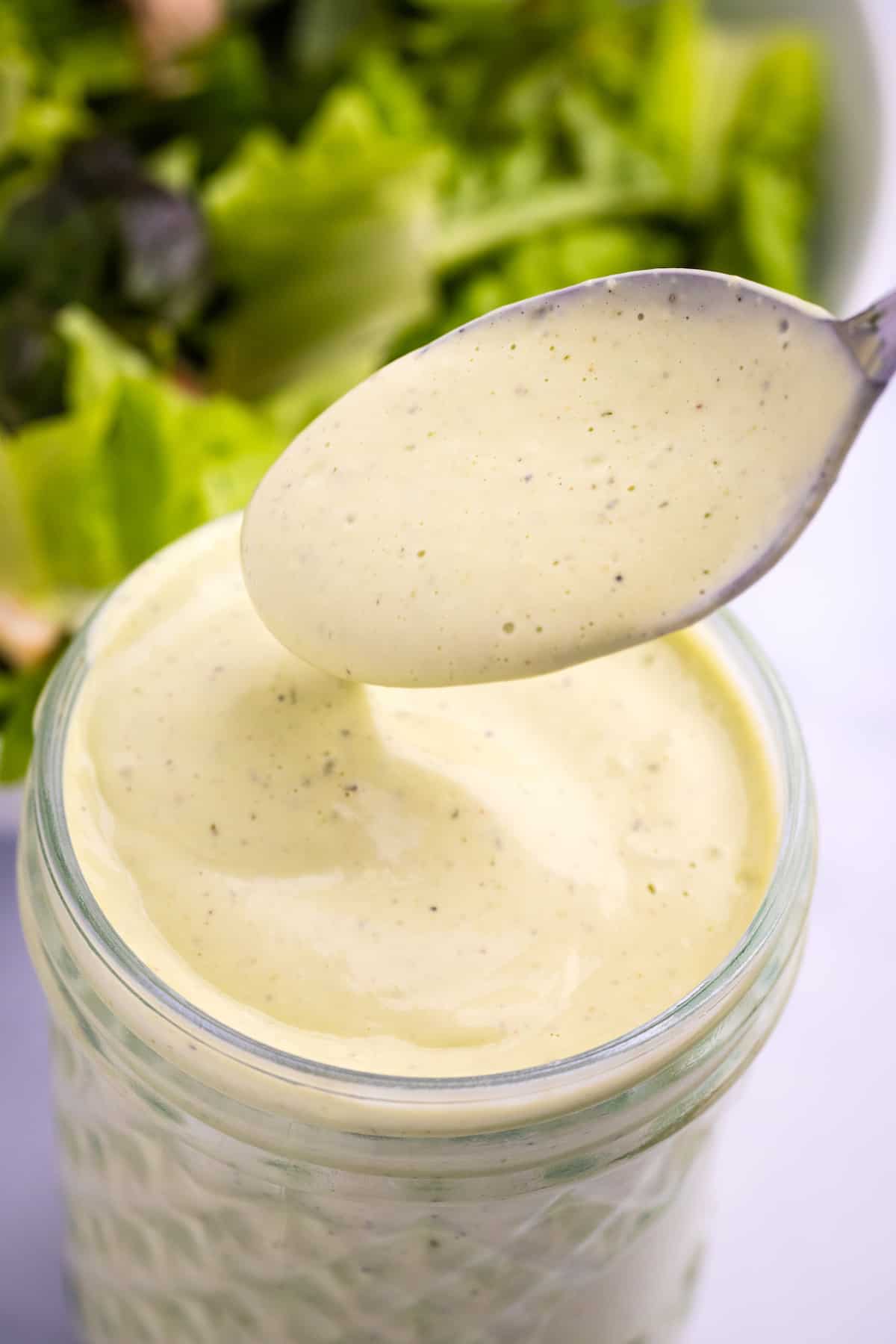 spoon and mason jar filled with avocado lime ranch dressing in front of a bowl of salad greens