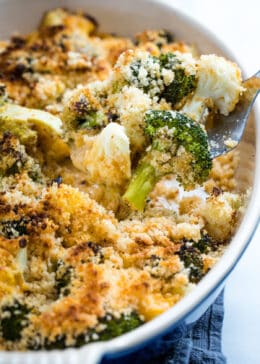 Broccoli and cauliflower in a cheesy sauce topped with breadcrumbs on a fork above the casserole dish.