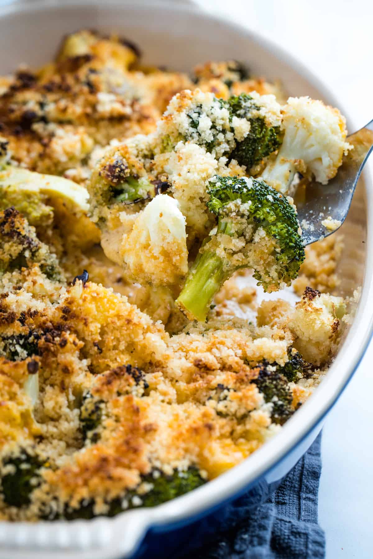 Broccoli and cauliflower in a cheesy sauce topped with breadcrumbs on a fork above the casserole dish.