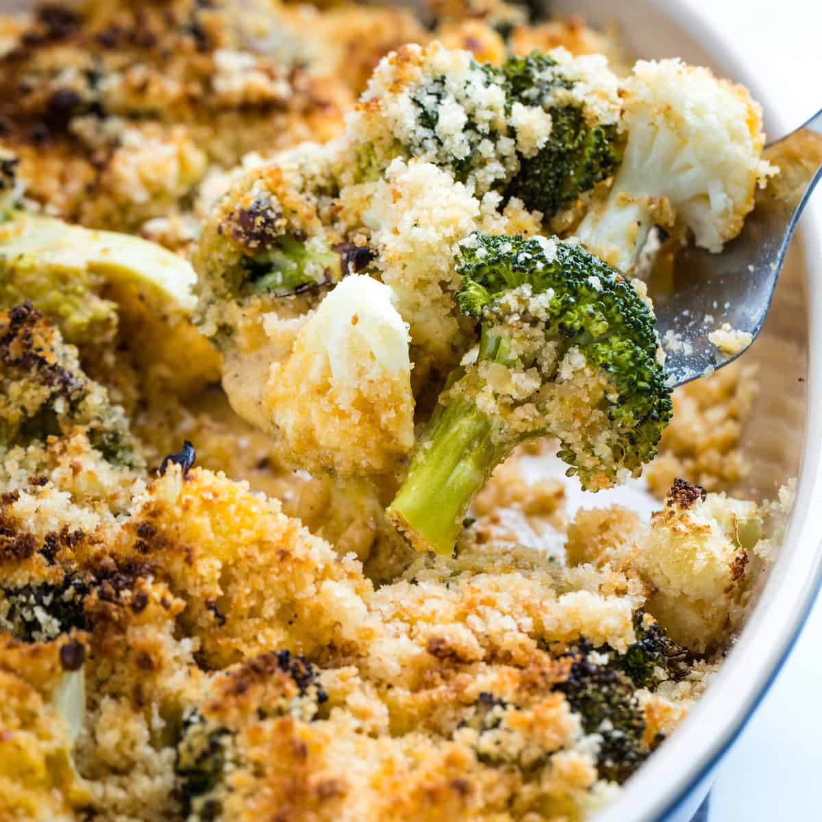 Broccoli and cauliflower in a cheesy sauce topped with breadcrumbs on a fork above the casserole dish.