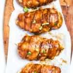 Four chicken breasts wrapped in bacon smothered in BBQ sauce sitting on parchment paper.