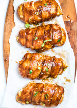 Four chicken breasts wrapped in bacon smothered in BBQ sauce sitting on parchment paper.