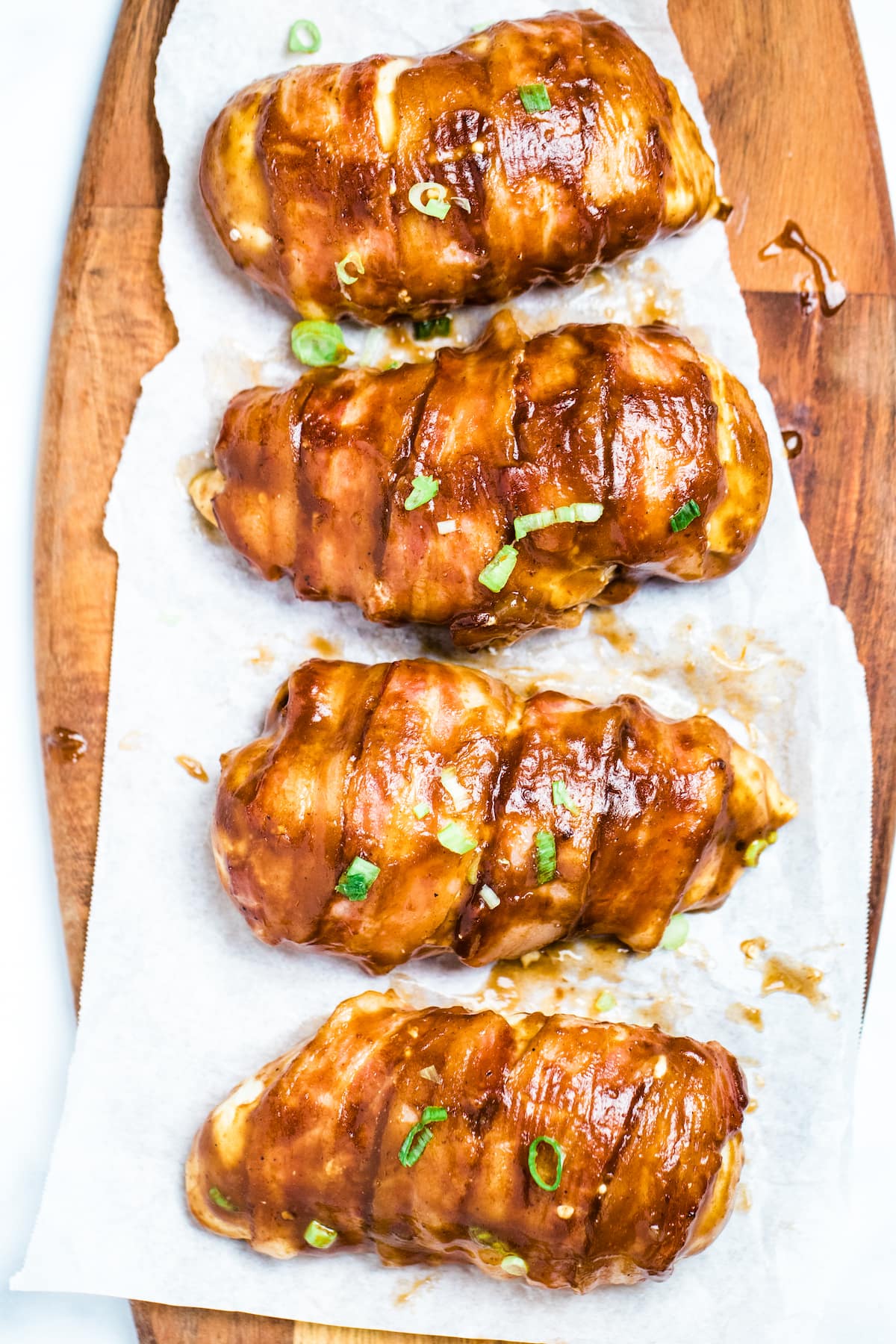 Four chicken breasts wrapped in bacon smothered in BBQ sauce sitting on parchment paper.