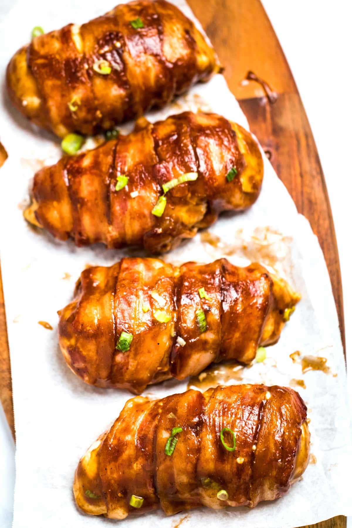 Four chicken breasts wrapped in bacon smothered in BBQ sauce sitting on parchment paper.