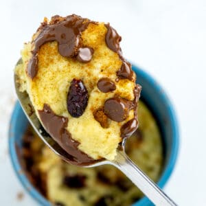 A spoon scooping out a piece of chocolate chip mug cookie from a blue cup.