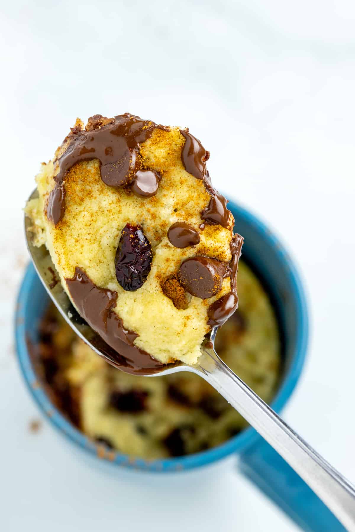 A spoon scooping out a piece of chocolate chip mug cookie from a blue cup.