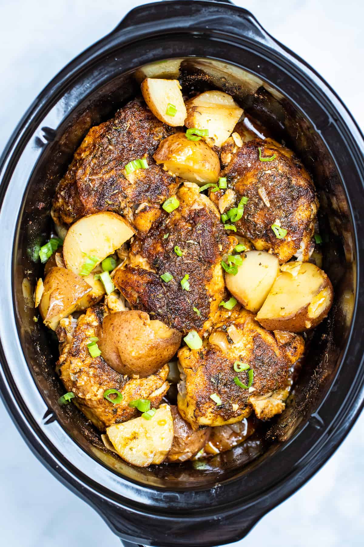 Slow cooker full of chicken thighs and potatoes topped with chopped green onions.