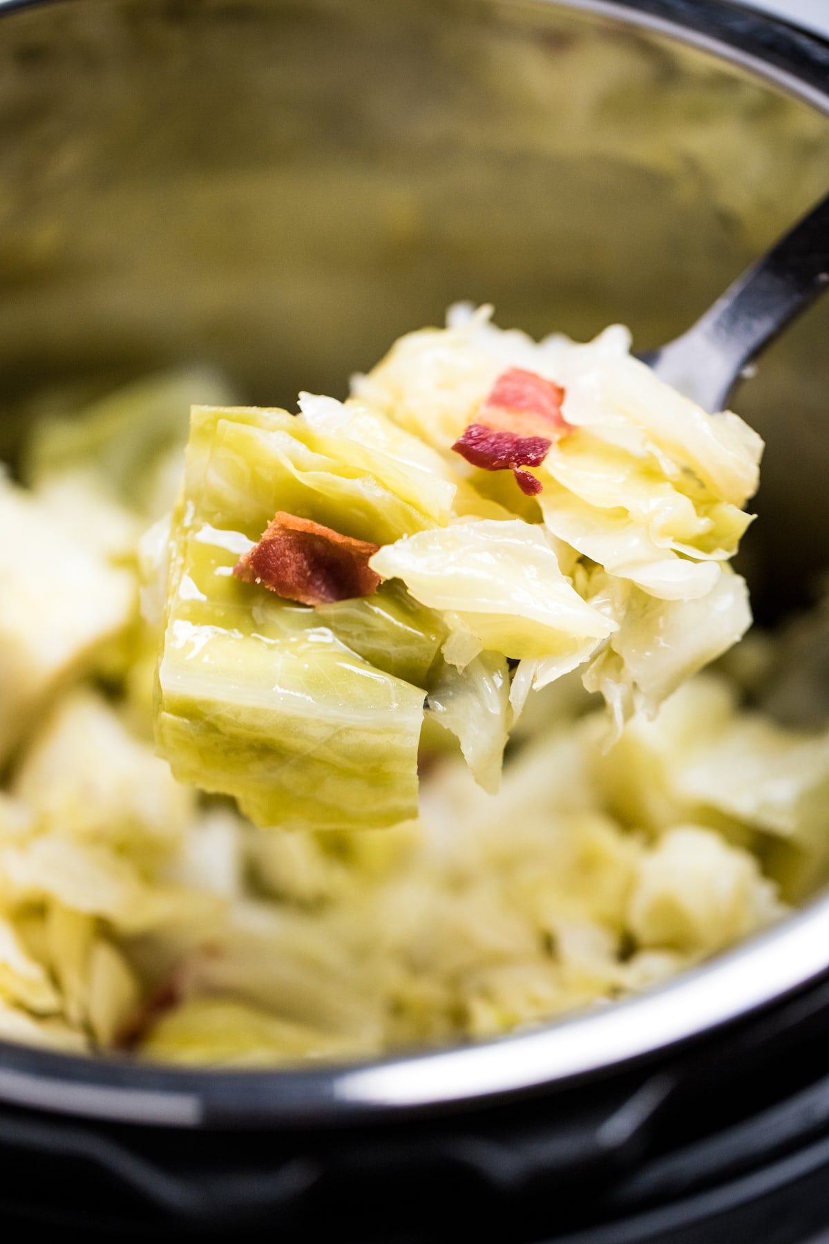 A spoon lifting a scoop of cooked cabbage topped with bacon pieces from an instant pot.