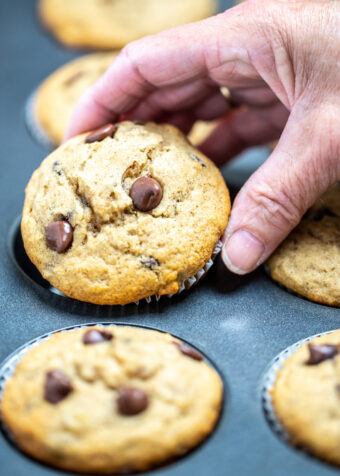A hand removing a chocolate chip banana muffin from a muffin tin.