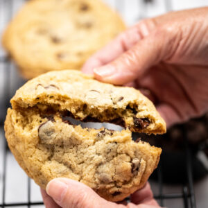 A chocolate chip cookie being broken in half by two hands.