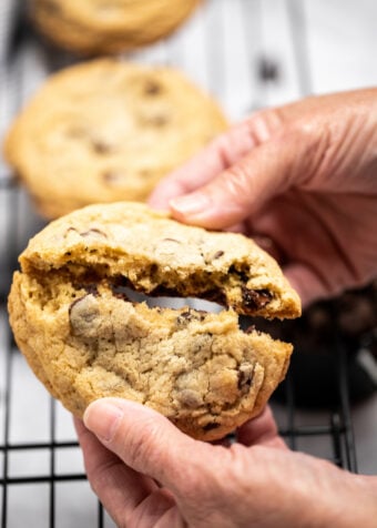 A chocolate chip cookie being broken in half by two hands.