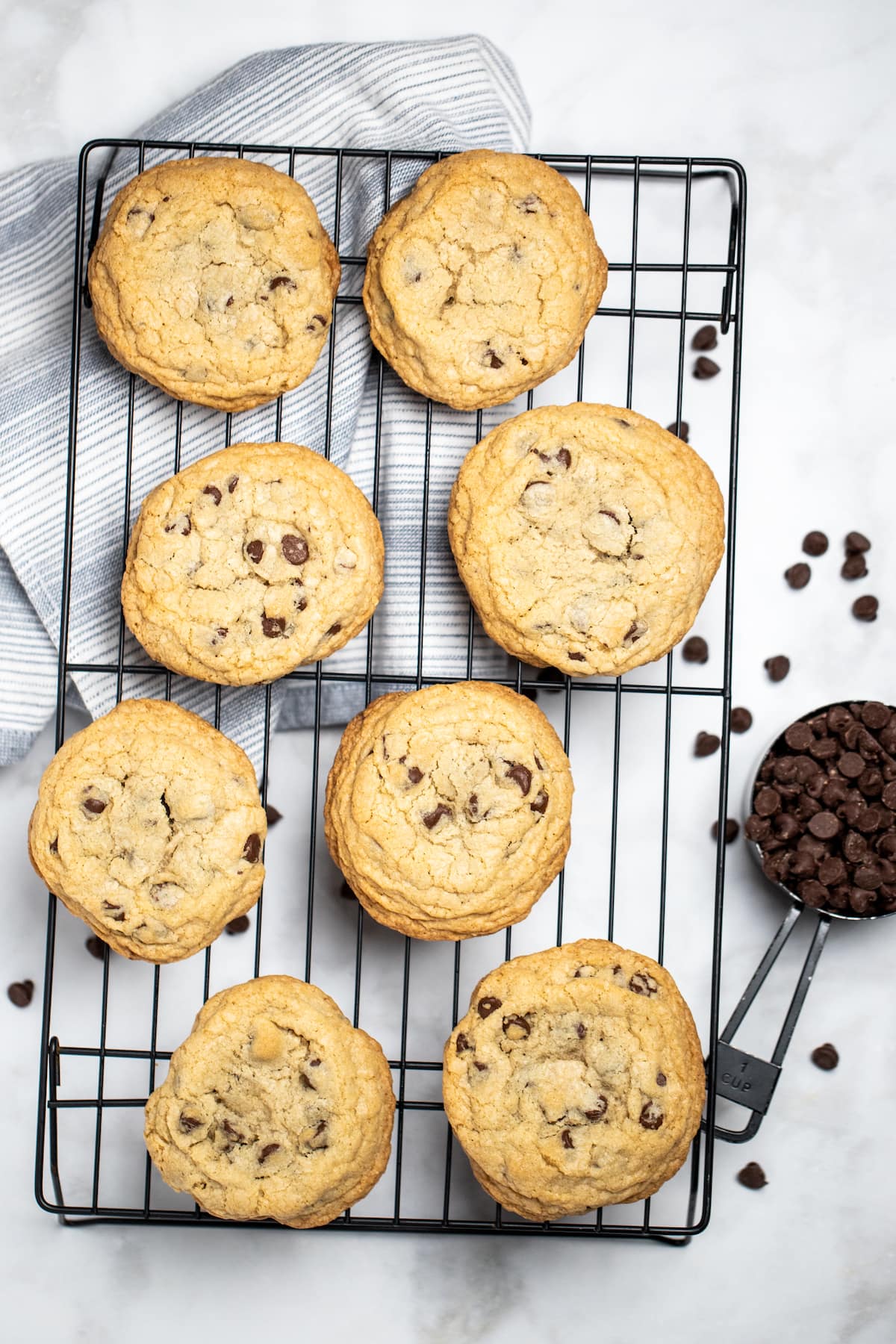 Large chocolate chip cookies sitting on a cooling rack with a measuring cup of chocolate chips next to it on a marble table.