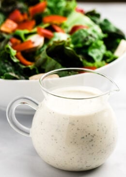 Small pitcher of buttermilk ranch dressing sitting in front of a bowl of salad.