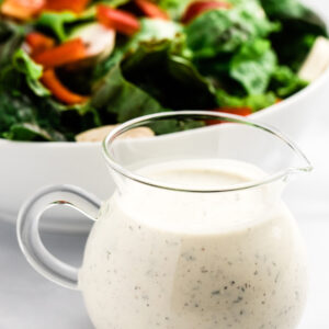 Small pitcher of buttermilk ranch dressing sitting in front of a bowl of salad.