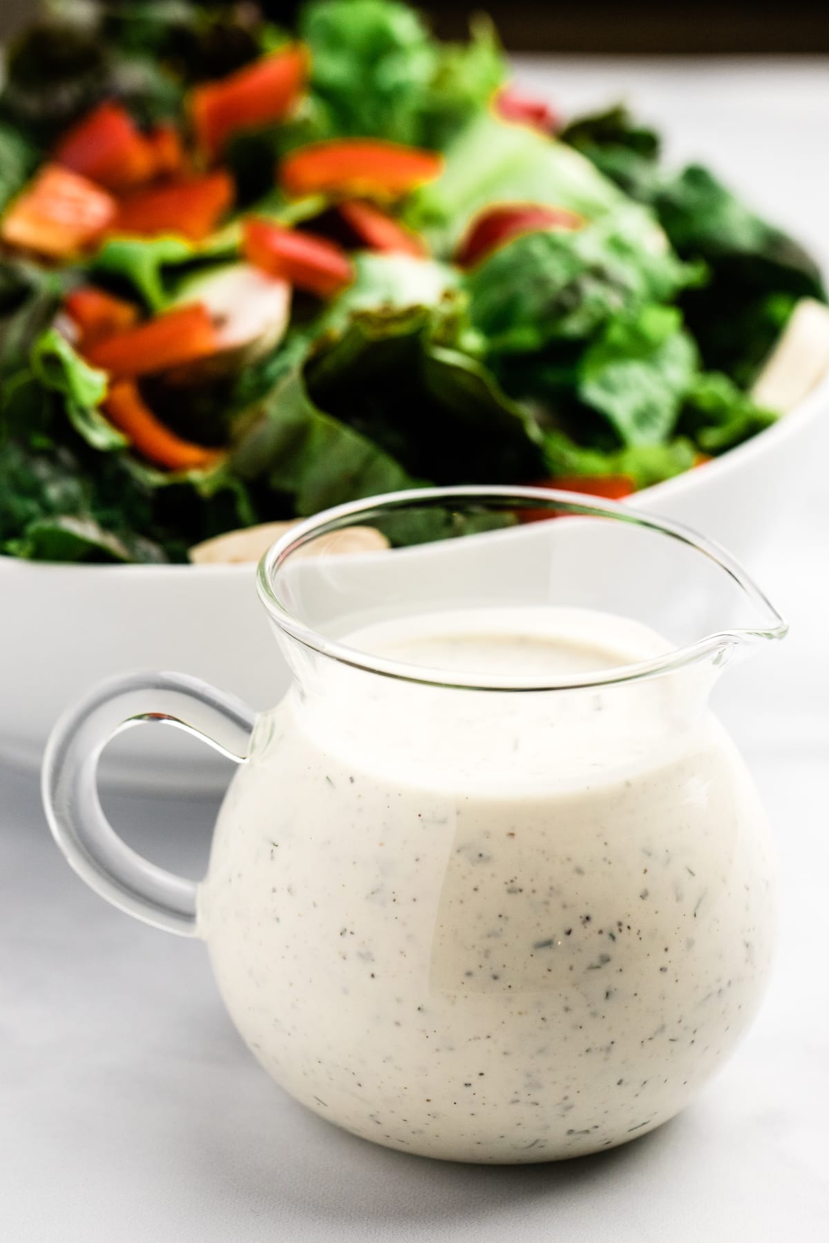 Small pitcher of buttermilk ranch dressing sitting in front of a bowl of salad.