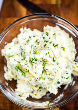 Herb butter in a clear glass bowl on a wooden cutting board.