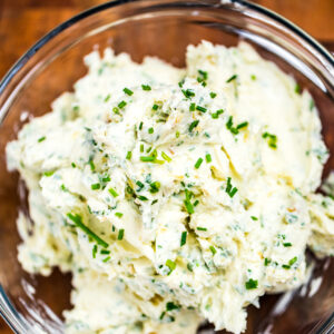 Herb butter in a clear glass bowl on a wooden cutting board.