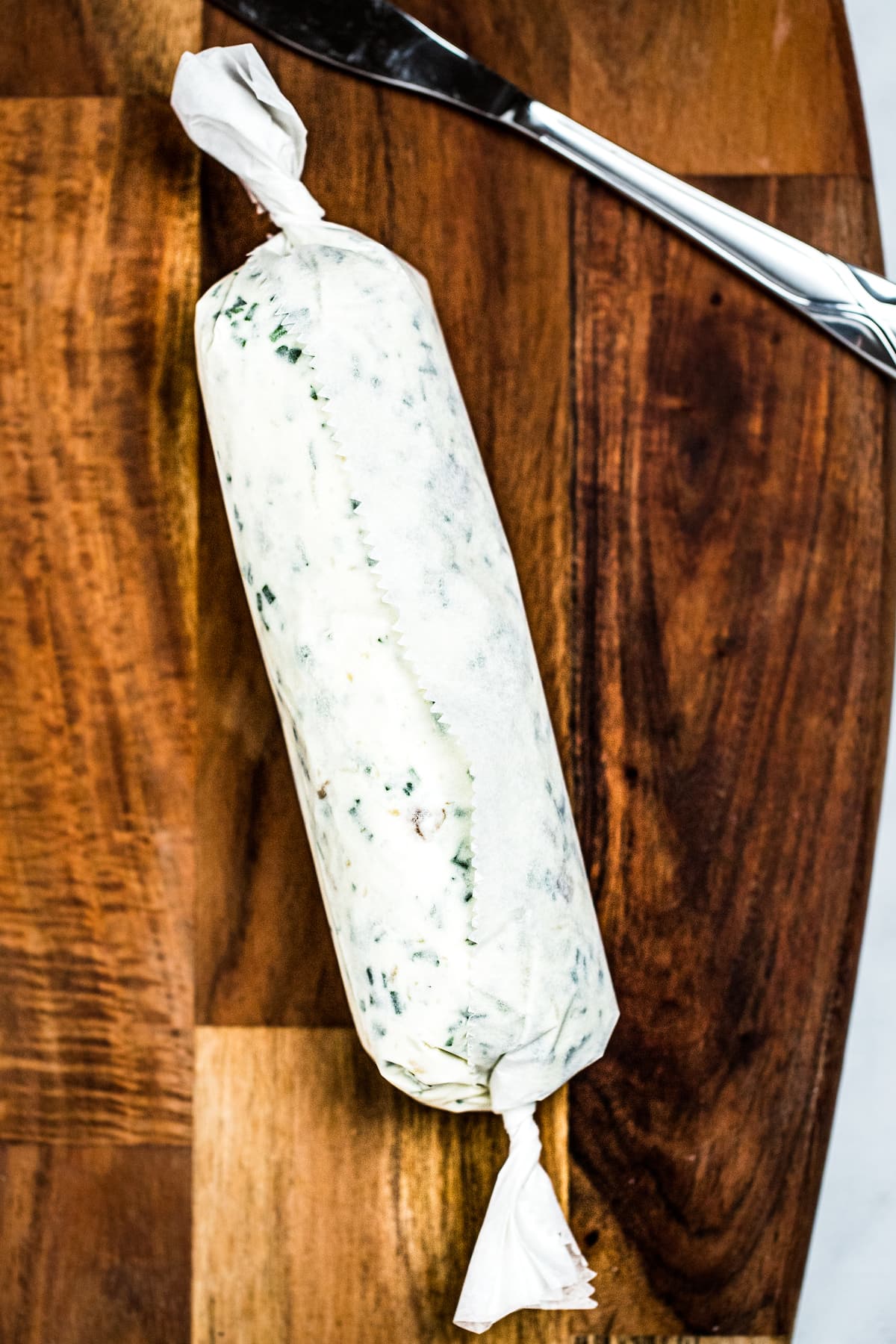 Herb butter wrapped into a log shape in parchment paper on a wooden cutting board next to a butter knife.