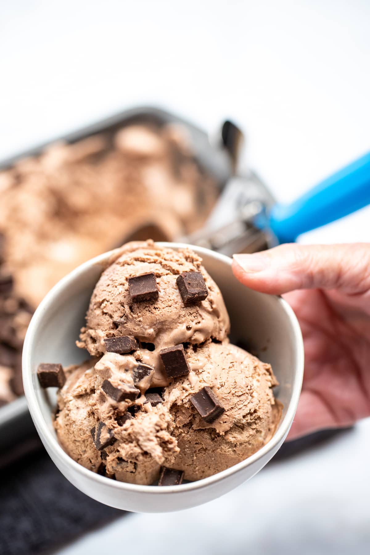 A hand holding a bowl of chocolate ice cream topped with chocolate chunks next to a bread pan of ice cream with an ice cream scoop.