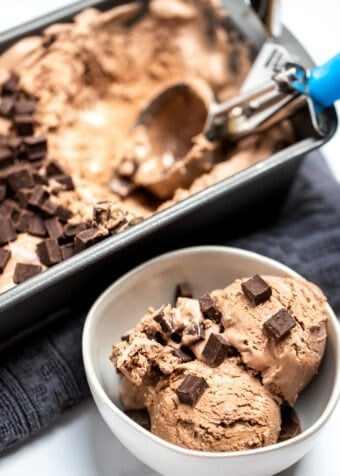 A bowl if chocolate ice cream topped with chocolate chunks next to a bread pan of ice cream with an ice cream scoop.