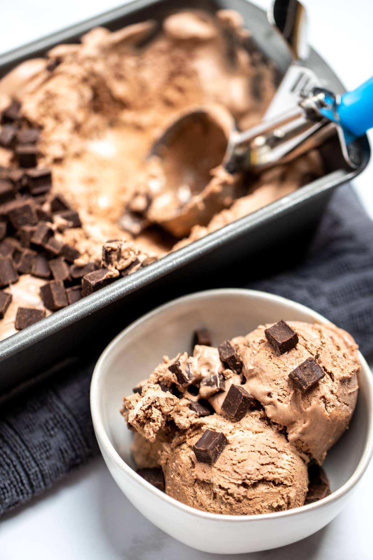 A bowl if chocolate ice cream topped with chocolate chunks next to a bread pan of ice cream with an ice cream scoop.