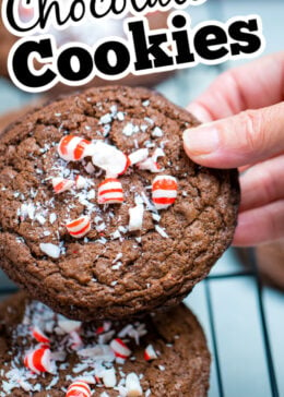A pinterest pin with a hand holding a peppermint chocolate cookie above other cookies on a cooling rack.