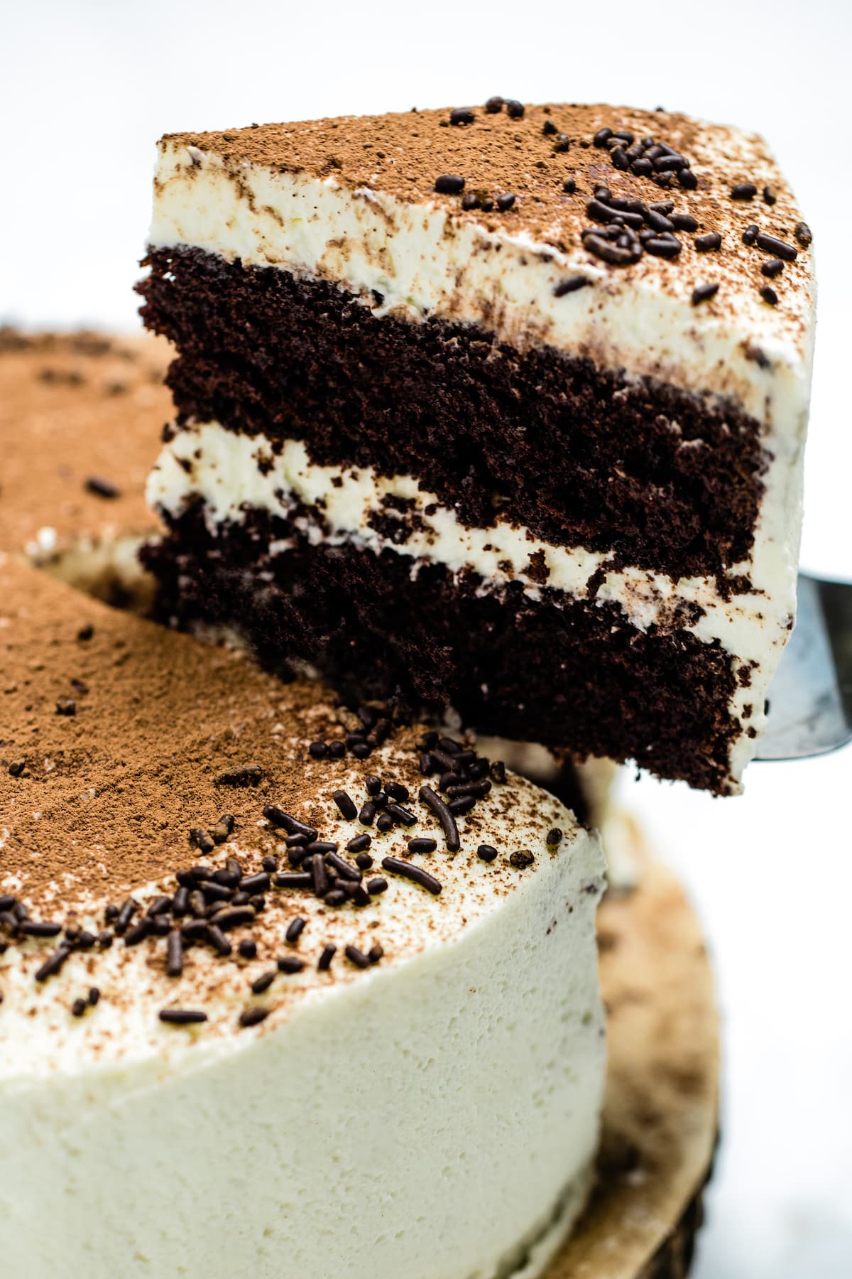 Chocolate coffee cake with a slice of cake being lifted up. The cake is coated in mascarpone frosting, with a middle layer of frosting, and decorated with chocolate sprinkles and a dusting of cocoa powder.