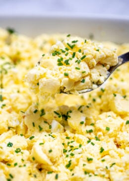 A spoon lifting scrambled eggs out of a baking dish topped with chives.