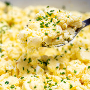 A spoon lifting scrambled eggs out of a baking dish topped with chives.
