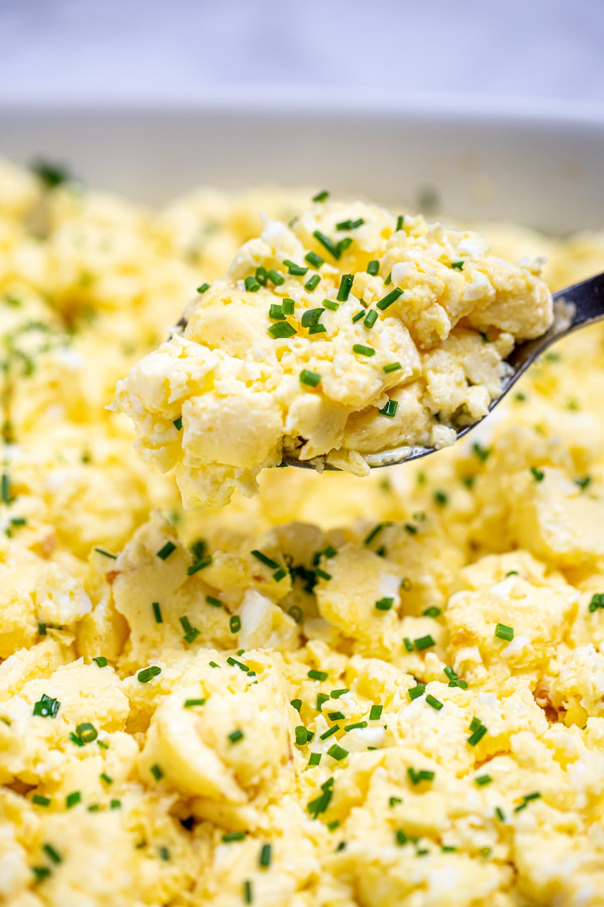 A spoon lifting scrambled eggs out of a baking dish topped with chives.