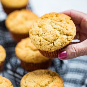 A hand holding a corn muffin above a cooling rack of other corn muffins.