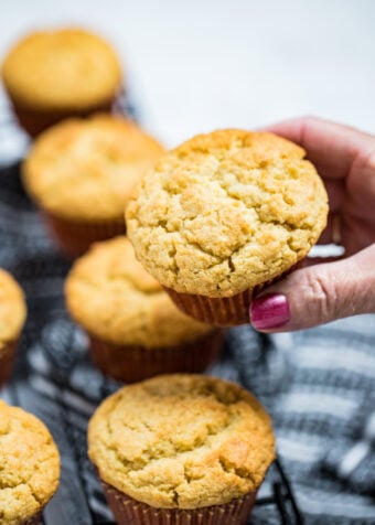 A hand holding a corn muffin above a cooling rack of other corn muffins.