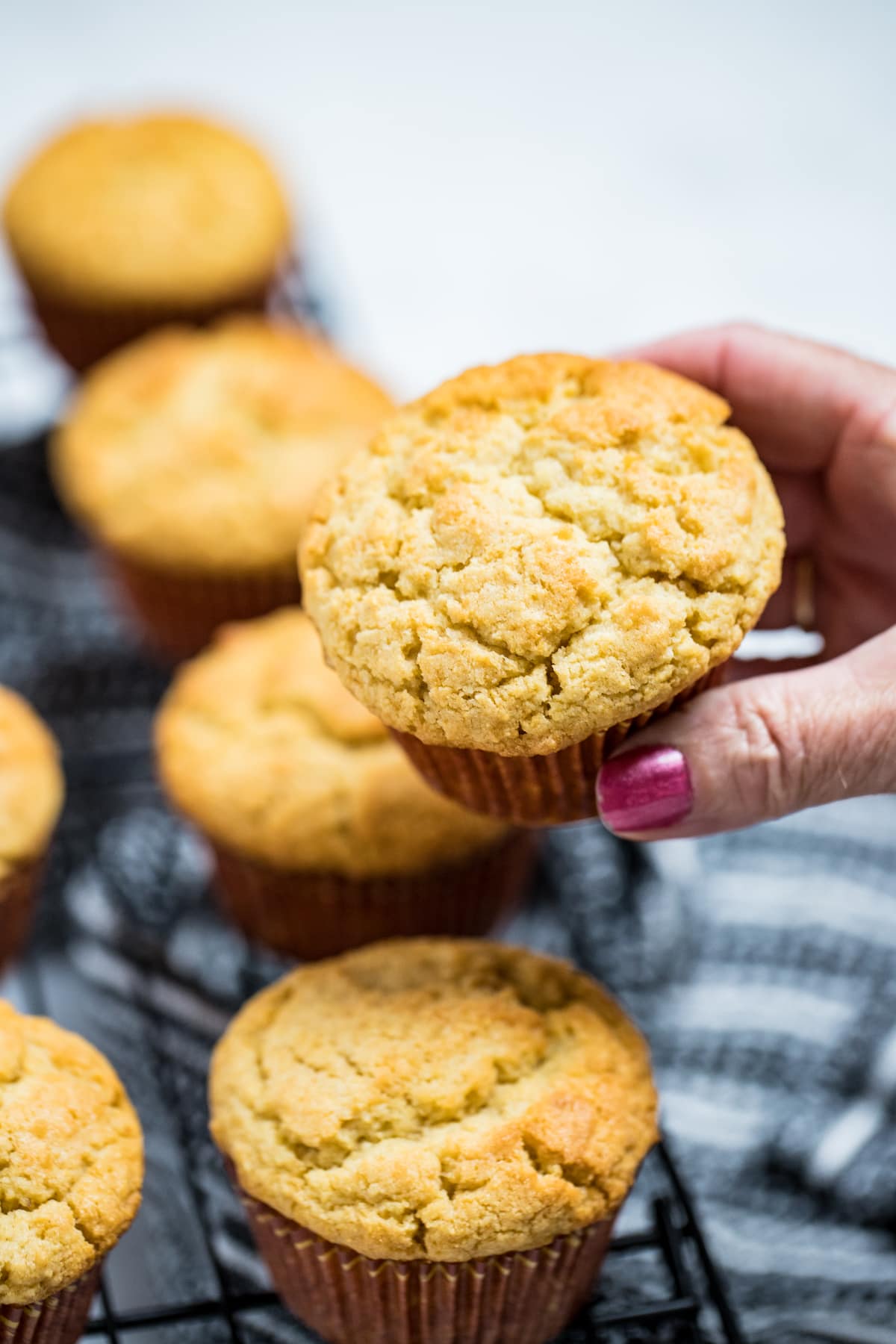 A hand holding a corn muffin above a cooling rack of other corn muffins.