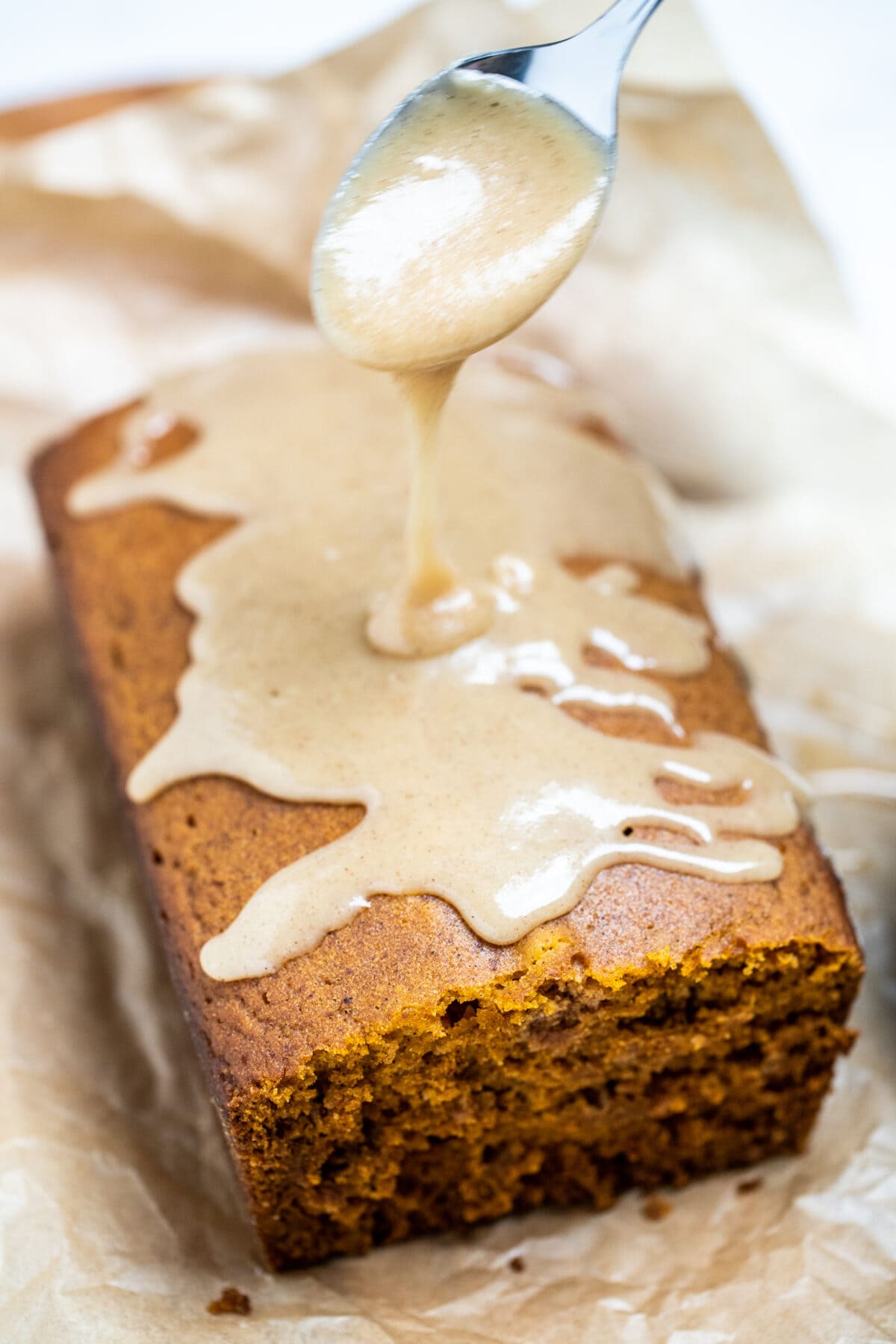 A loaf of pumpkin bread on a piece of parchment paper with a spoon drizzling pumpkin spice glaze on top.