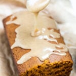 A loaf of pumpkin bread on a piece of parchment paper with a spoon drizzling pumpkin spice glaze on top.