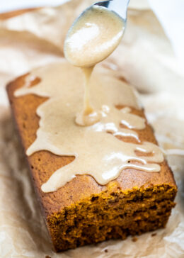 A loaf of pumpkin bread on a piece of parchment paper with a spoon drizzling pumpkin spice glaze on top.