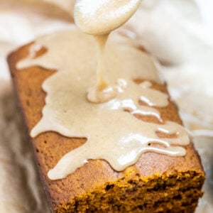 A loaf of pumpkin bread on a piece of parchment paper with a spoon drizzling pumpkin spice glaze on top.