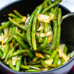 A bowl of instant pot green beans on the table topped with shredded parmesan cheese, and a spoon lifting out a serving of beans.