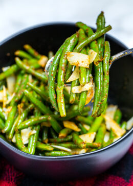 A bowl of instant pot green beans on the table topped with shredded parmesan cheese, and a spoon lifting out a serving of beans.