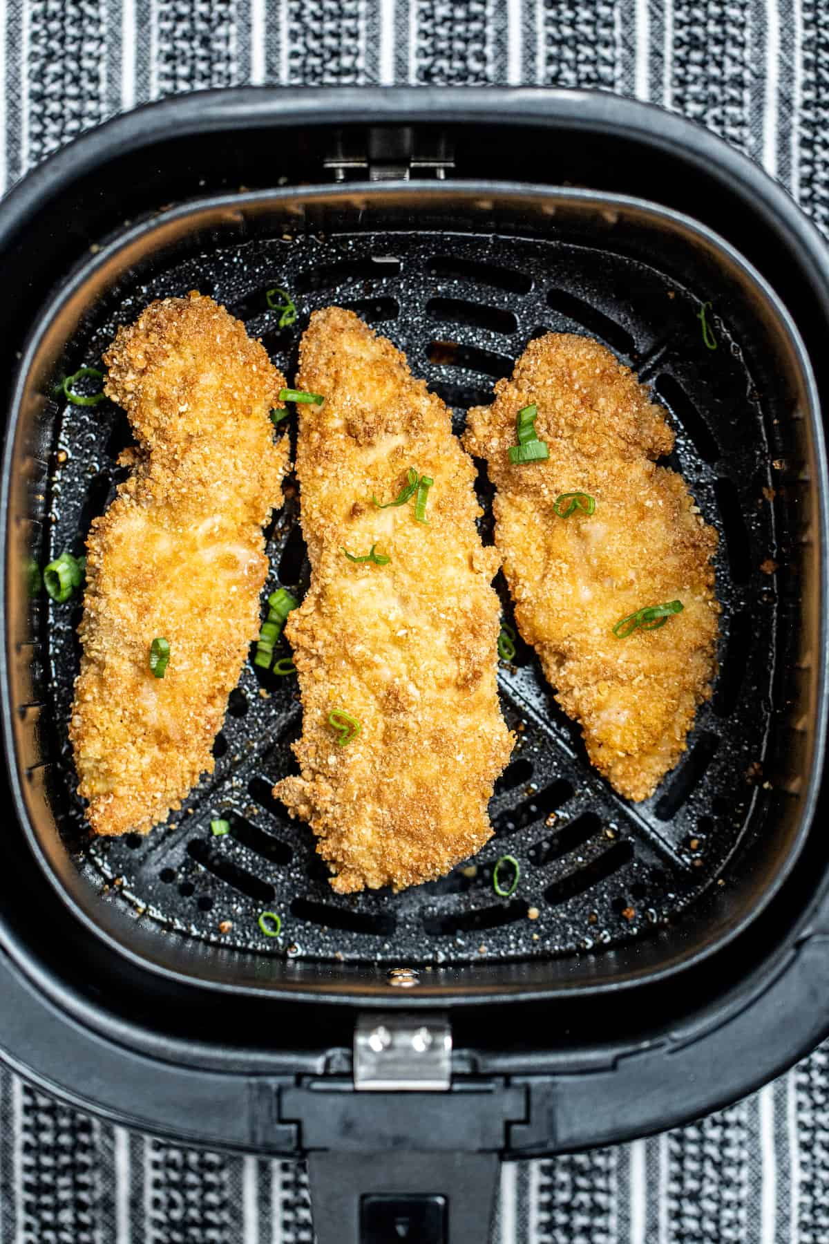 Air fryer chicken tenders in the air fryer basket topped with green onion pieces.